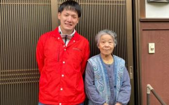 赤い制服を着た男性スタッフが高齢女性と一緒に玄関前で笑顔で撮影した写真